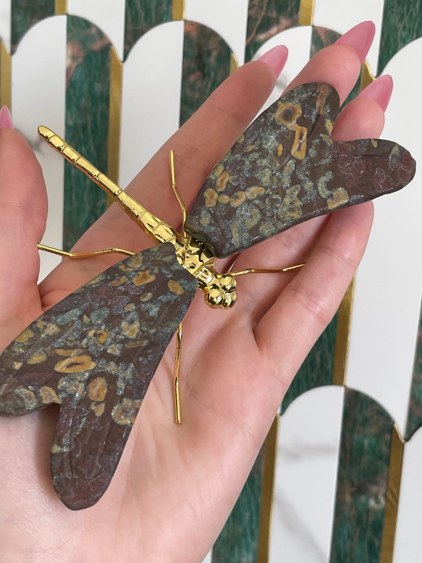 Decorative dragonfly with gold and bronze details held by a hand against a patterned background